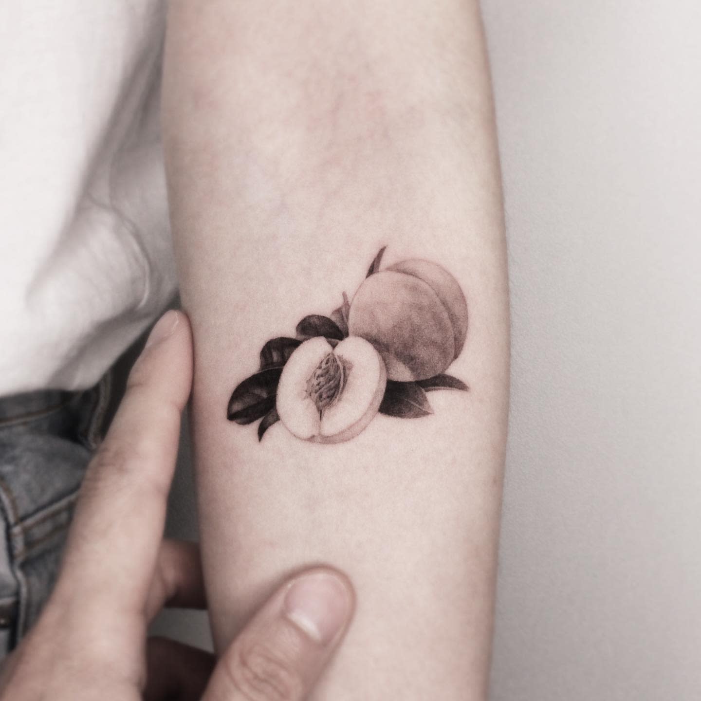 Delicate black and gray tattoo of a realistic peach with a sliced half and leaves on the arm